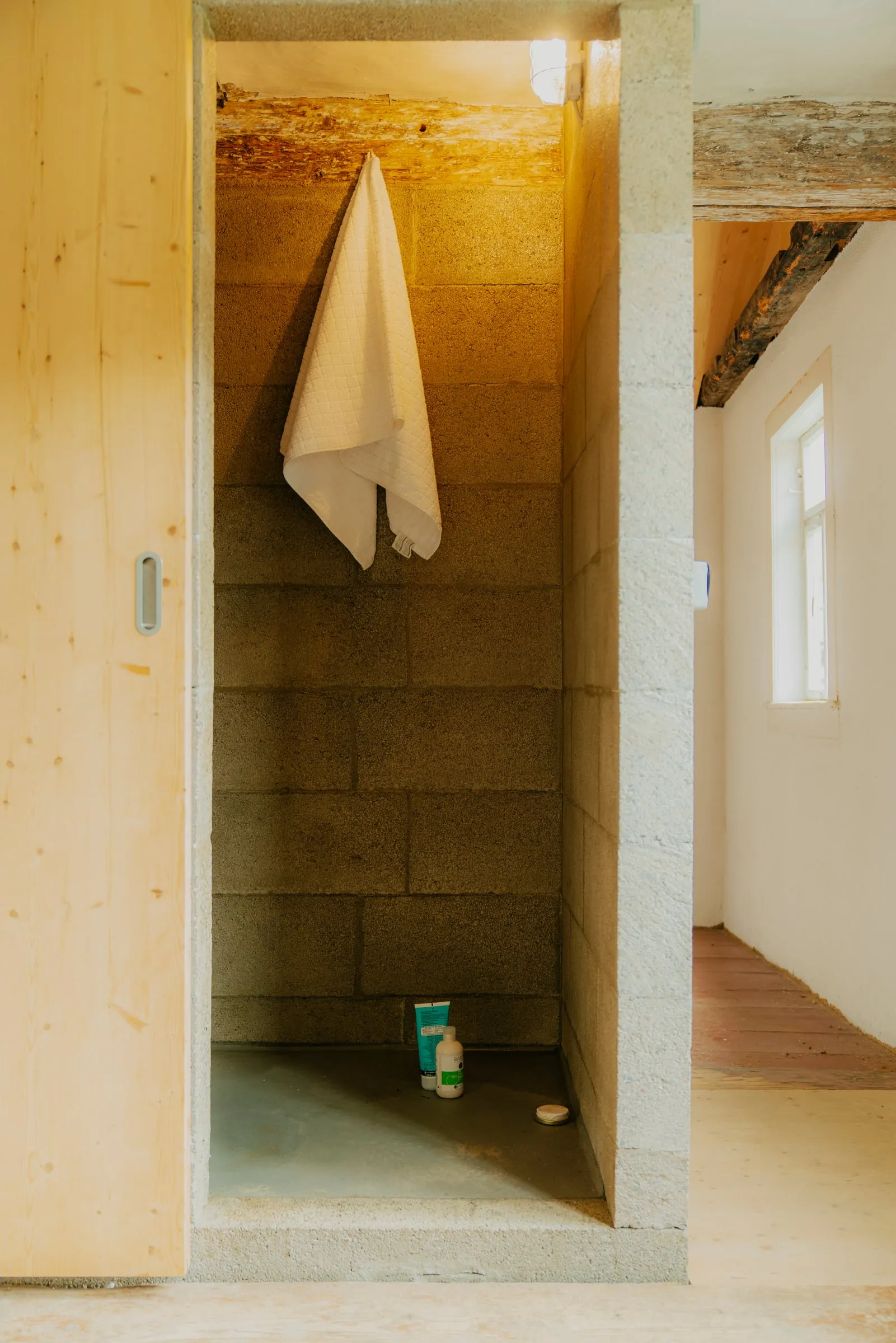 Dusche aus Betonsteinen hinter einer Schiebetür aus Holz, an der Wand hängt ein Handtuch