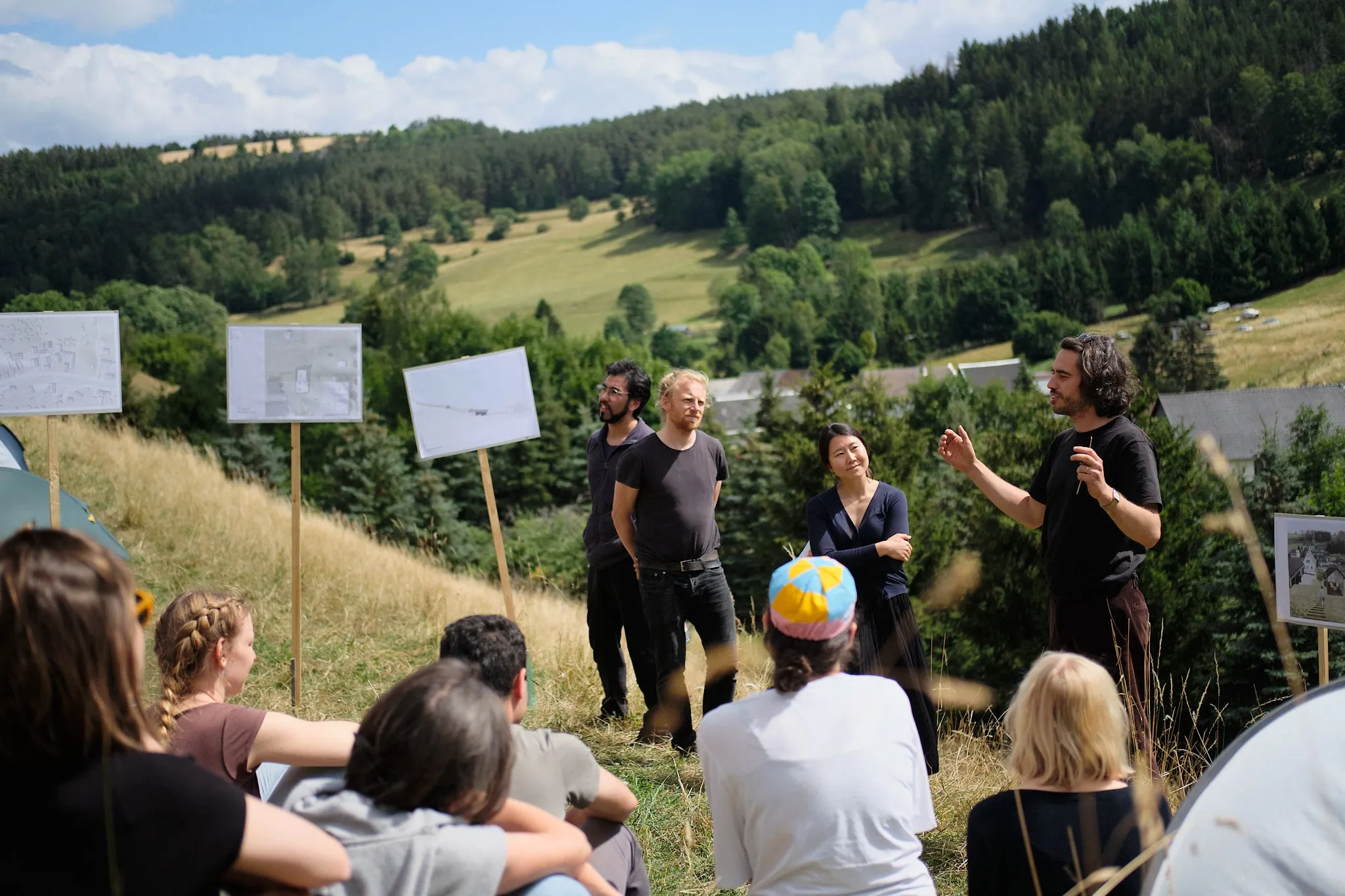 Studierende sitzen auf einer Wiese am Hang, vor ihnen stellen vier Personen neben Plakaten auf Holzstäben ihre Arbeit vor