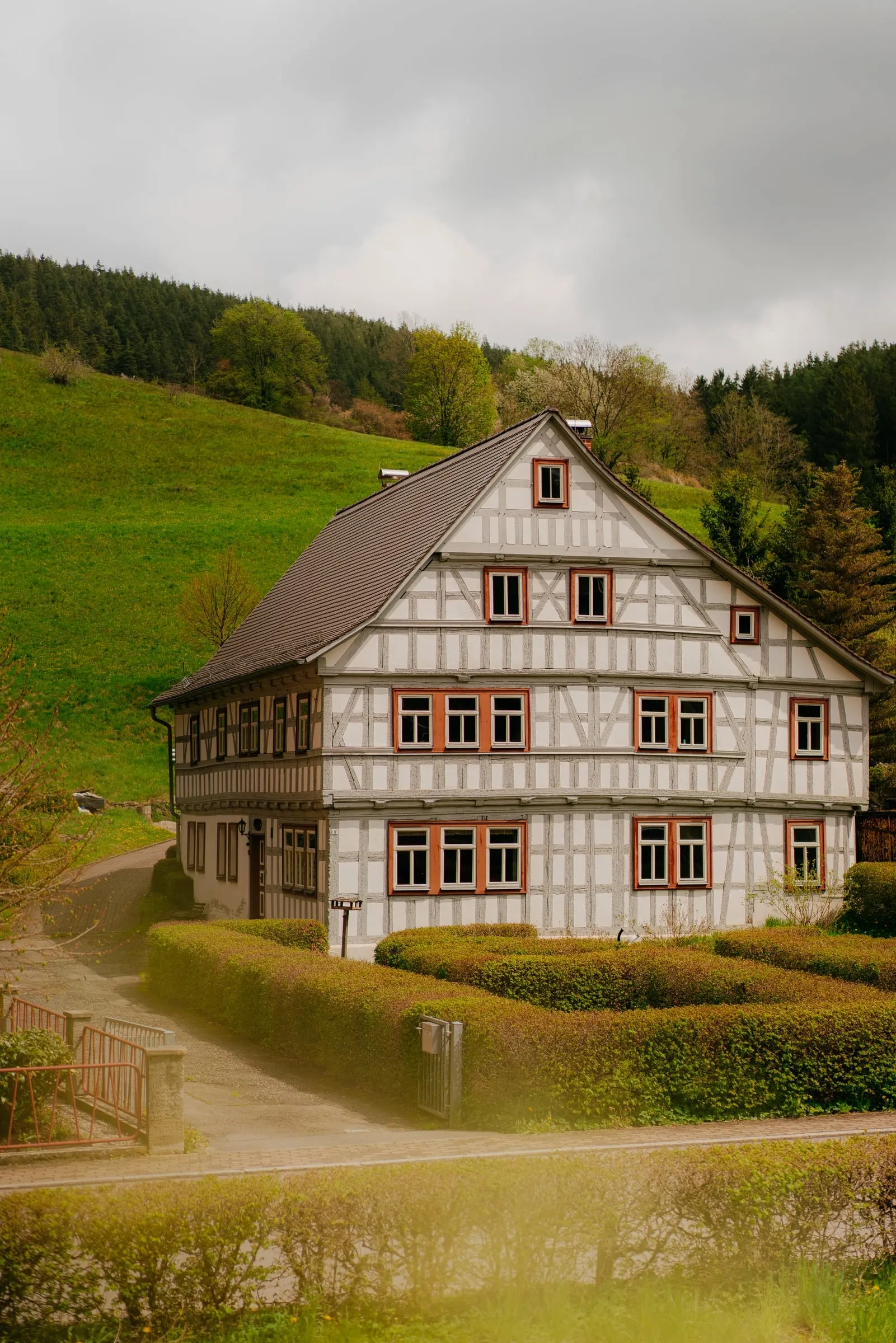 Weißes Fachwerkhaus mit Satteldach und roten Fensterrahmen vor einer grünen Hangwiese, davor Hecken und die Dorfstraße