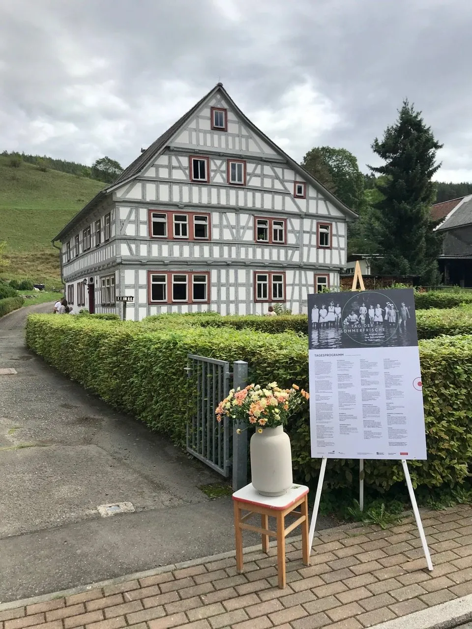 Das Haus am Tag der Sommerfrische, davor eine Staffelei mit Informationstafel und ein Blumenstrauß
