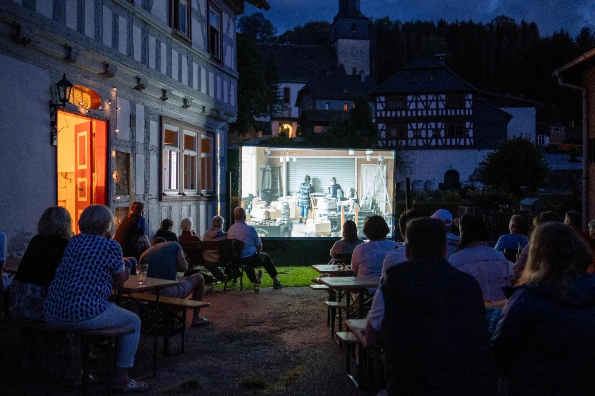 Abends sitzen Gäste an Biertischen vor dem Haus und schauen auf eine Leinwand, im Hintergrund Fachwerkhäuser und der Kirchturm