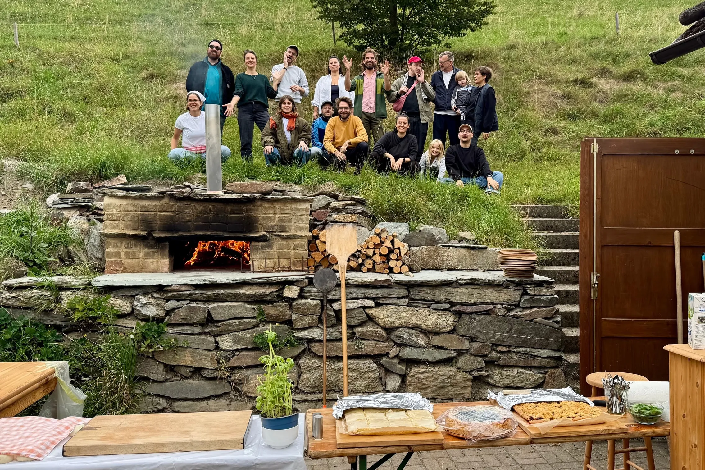 Gruppenfoto am Hang hinter dem Haus, davor der Pizzaofen aus Lehm mit Feuer und ein Tisch mit Kuchen