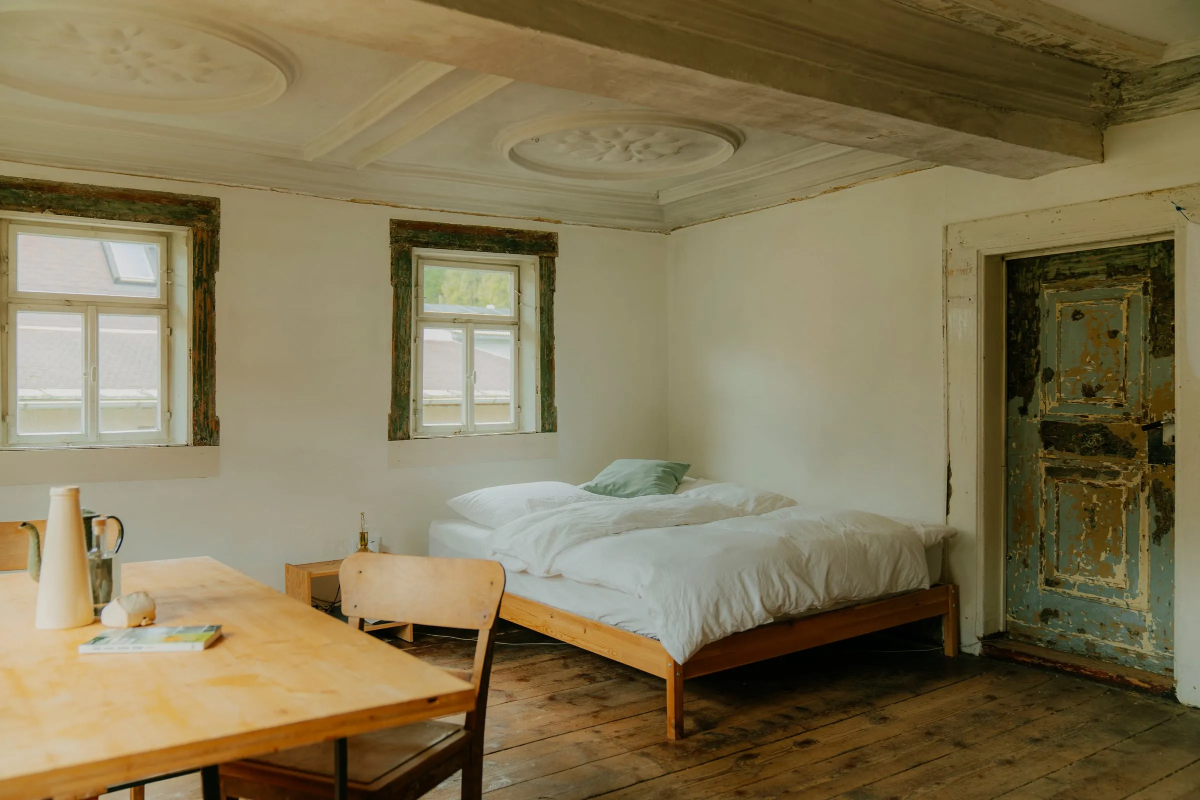 Schlafraum mit Stuckdecke, Doppelbett, Holztisch und einer alten, abgeblätterten Tür